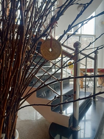 Foreground: A dense arrangement of brown, budding branches with small cotton-like buds. A golden, circular coin with a square hole hangs from one branch with a red string.
Background: A staircase with black treads and a silver railing ascends. Beyond the staircase is a large window letting in daylight. A few tables and chairs are partially visible in the room beyond the window.