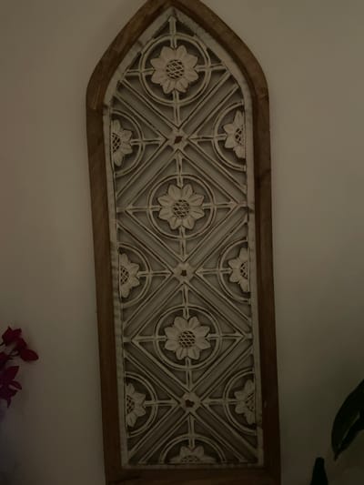 The image shows a decorative wall hanging with a wooden frame. The hanging has an elongated, arrow-like shape with a pointed end on the left. Within the frame, there is a white metal lattice pattern featuring repeating floral designs arranged in circular and diamond shapes. The flowers have a central circular detail with petal-like projections extending outwards. The background is a light beige or cream color, likely a wall. In the bottom right corner, there are a few red and pink flowers or leaves, possibly from a houseplant. The lighting appears soft and artificial. The location is unspecified, and there is no indication it is in Bengaluru, India. There are no people visible in the image.