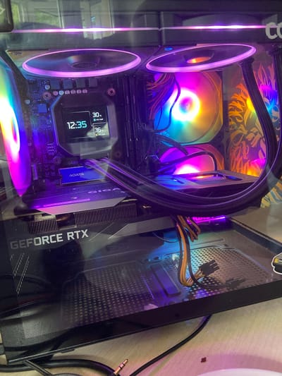 The image shows the interior of a computer tower, likely a gaming PC, with visible components illuminated by RGB lighting. A central liquid cooler with RGB fans is prominently displayed, with one fan showing a rainbow spectrum of colors and the other a pinkish-purple hue. The motherboard is visible with RAM sticks labeled "FURY" and an ADATA SSD. A graphics card with "GEFORCE RTX" branding is installed below the motherboard. A small digital display on the CPU cooler shows the time as 12:35 and CPU temperatures of 30°C and 36°C. The computer case appears to have a glass side panel, allowing for a clear view of the internal components. Cables, including power connectors, are neatly routed. The background is blurred, but appears to be an indoor setting with a wooden surface. There are no people or animals present in the image. The location of Beloretsk, Russia is not discernible from the image itself.