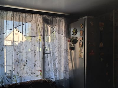 The image depicts an interior scene, likely a kitchen or dining area, in Kremenchuk, Ukraine. A large window is covered by sheer, floral-patterned curtains, through which a building and trees are visible. To the right of the window, a stainless steel refrigerator stands against a wall. The refrigerator door is adorned with numerous magnets, some of which appear to be decorative or souvenir items, including what looks like an Orthodox icon and some Cyrillic lettering. The lighting suggests it is daytime, with sunlight streaming through the window, casting shadows across the room and illuminating parts of the refrigerator. A portion of a patterned sofa or chair is visible in the lower left corner of the frame. The overall environment appears to be a domestic setting.