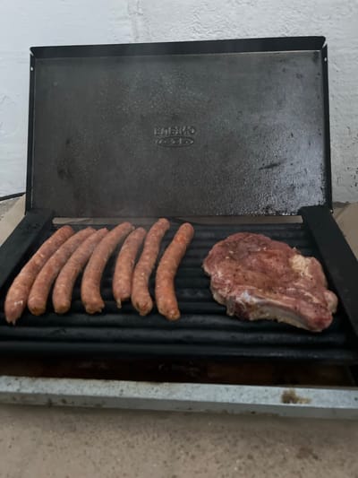 The image depicts an outdoor grilling scene, possibly in Vratsa, Bulgaria, given the context of the request. A black grill, with the brand name "EDAKO" visible on its upper lid, is open and positioned outdoors. On the grill grates, there are seven sausages laid out in a row and a single piece of seasoned meat, likely a pork chop or steak, placed to the right of the sausages. The grill appears to be heated, as indicated by faint wisps of smoke rising from the food. The background consists of a white, textured wall, possibly brick, and the ground beneath the grill is a concrete or stone surface. The overall scene suggests a preparation for or the early stages of cooking a meal outdoors.