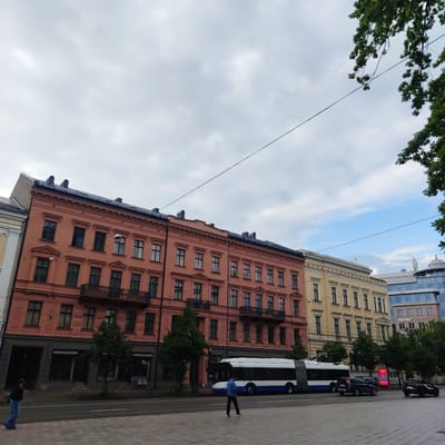 The image captures a street scene in Riga, Latvia, likely near the city center, with a line of historical buildings on the left and a modern glass building on the right. The buildings on the left appear to be old, with the closest one painted in a salmon pink color and featuring ornate architectural details. The building next to it is yellow with classical elements. Several trees are visible along the street.

In the foreground, a wide pedestrian walkway is paved with patterned tiles. A street runs alongside the walkway, with a trolleybus and several cars on it. The trolleybus is white with blue accents and has its poles extended to connect to overhead wires. Two individuals are visible on the walkway: one person in a dark jacket and light-colored pants walks away from the camera, and another person in a blue denim jacket and dark pants walks towards the right of the frame, looking at their phone. A third person, dressed in a light blue jacket and dark pants, is walking across the street in front of the trolleybus.

The sky is partly cloudy, with patches of blue visible. The overall impression is of an urban environment with a mix of old and new architecture, a common sight in European cities. The specific location, given the architecture and the presence of a trolleybus, is characteristic of Riga's central districts. The "KALVE" signage visible on the storefronts of the salmon-pink building suggests a commercial area.