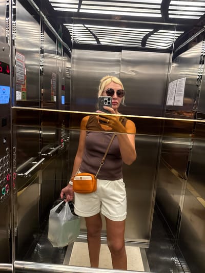 A woman stands in an elevator, taking a selfie. She has blonde hair and is wearing sunglasses, a brown sleeveless top, and white shorts. She is carrying a clear plastic bag in her left hand and a small orange and white crossbody bag. The elevator walls are mirrored stainless steel, reflecting the overhead lights and other parts of the elevator. There are elevator buttons and a digital display visible on the left wall, showing a red arrow pointing down and the number "008". The floor of the elevator appears to be tiled. The scene is consistent with being in an elevator in Alanya, Turkey, a coastal city on the Mediterranean coast of Turkey known for its tourist accommodations.