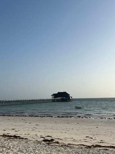 This image, set in Kiwengwa, Tanzania, depicts a tranquil coastal landscape under a vast, clear sky. The sky transitions from a soft blue on the left to a lighter, almost hazy white towards the right horizon, indicative of daytime, possibly late morning or early afternoon, with a bright sun.
The central feature is a distinct building constructed on stilts, situated over calm, shallow turquoise waters. The building has a dark, possibly thatched roof and lighter-colored walls with blue details, appearing to be an overwater restaurant or resort structure. A long, narrow wooden pier extends from the left, providing access to this building, its supports visible against the water.
To the right of the water and pier, a sandy beach runs vertically through the frame (when correctly oriented), characterized by light-colored sand interspersed with natural dark patches of seaweed or debris, particularly along the waterline. A small, simple boat is visible floating peacefully in the water near the overwater building. The water surface shows gentle ripples, reflecting the calm conditions. No people are present, enhancing the serene and untouched quality of the scene. No text is visible in the image.