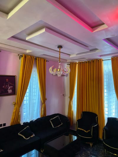The image displays a modern interior living space situated in Alor, Nigeria. The room features a multi-tiered false ceiling with integrated linear lighting elements emitting pink and red light. A gold-finished chandelier with five crystal-decorated, curved arms hangs centrally. Light-colored walls are flanked by several windows covered with floor-length, golden-yellow curtains secured by gold tassels, revealing sheer white inner drapes. Furniture includes a black sectional sofa with black and gold-trimmed throw pillows, positioned on the left, and at least two black armchairs with gold accent trim and gold-colored legs on the right. A dark, reflective coffee table occupies the central foreground. The floor is covered in dark, reflective tiles. A framed portrait depicting a young boy holding an item resembling a book or tablet hangs on the left wall. An air conditioning unit is mounted on the wall to the right of the chandelier. The scene is devoid of people, actions, or interactions.