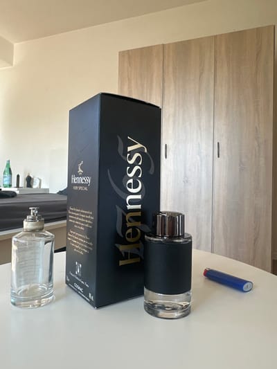 The scene is an interior room in Höri, Switzerland, presented with a forensic perspective. In the foreground, positioned on a light-colored, possibly white, flat surface, are three primary objects arranged from left to right: an empty, clear glass bottle with an octagonal base, fitted with a silver-colored pump dispenser and a beige collar; a tall, black rectangular cardboard box labeled "Hennessy VERY SPECIAL" with gold and white text, prominently displaying the brand logo and indicating "COGNAC" and "40% vol"; and a cylindrical glass bottle containing a clear liquid, featuring a dark, textured band around its middle and topped with a reflective silver cap. To the far right, partially visible on the same surface, is a blue disposable lighter.
The background consists of light-colored walls and large, light-wood-grain wardrobes with three visible doors, each equipped with a vertical dark handle. To the far left background, a dark-sheeted bed is partially visible, and on a surface behind it, a green bottle (possibly San Pellegrino) and several dark, slender objects are discernible. No individuals are present, and no active interactions are depicted. The environment is uniformly lit.