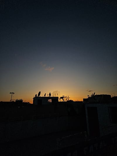 A panoramic, low-light photograph captures the urban landscape of Fez, Morocco, during a sunset or twilight period. The upper sky is a deep indigo, gradually transitioning to an orange-yellow glow along the horizon, punctuated by a solitary, wispy cloud. The city's structures are rendered in dark silhouette, dominating the lower two-thirds of the frame. Multiple parabolic satellite dishes, various antennas, and utility structures are distinct on numerous flat rooftops across the midground. Buildings feature dark openings suggestive of windows or doorways. The extreme foreground contains a dark, indeterminate structure bearing faint, partially legible white text, appearing to read "FATAL." No human figures are present.
