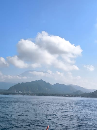 The image depicts a serene coastal landscape with a body of water in the foreground and a mountainous coastline in the background. The water is a deep blue, with gentle ripples suggesting calm conditions. In the distance, a range of lush green mountains forms the coastline, dotted with scattered buildings and vegetation. The sky above is a vibrant blue, partially obscured by large, fluffy white clouds. The overall impression is one of natural beauty and tranquility. The scene is consistent with views found in Karangasem, Bali, Indonesia, known for its picturesque coastlines and volcanic mountains.
