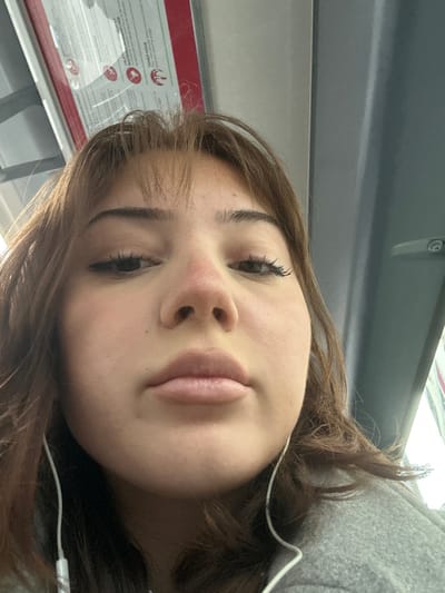 A close-up, low-angle self-portrait features a female individual with light brown hair and bangs, positioned within what appears to be the interior of a public transport vehicle in Schiltigheim, France. Her eyes are directed downwards towards the camera, and her lips are closed. The bridge of her nose exhibits visible redness. She is wearing white wired earphones, with the wires visible extending from beneath her chin. A grey garment, possibly a coat or hoodie, is visible on her left shoulder. In the upper left background, an overhead red and white sign with French text is partially visible. To the upper right, a grey vertical panel with a light-colored handle indicates the vehicle's interior structure.