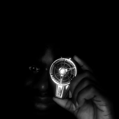 The image is a close-up, black and white photograph featuring a hand holding a metallic object. The hand, dark in tone, is positioned on the right side of the frame, gripping the object. The metallic object appears to be circular with a central design resembling a fan or propeller, reflecting light, suggesting a glossy surface.
Below the object, a reflection of a face is visible on a shiny surface. The eye of the person is visible and looks up towards the object in the hand. The background is entirely black, suggesting a dark environment or a focused spotlight on the subject.
There is no information to confirm that this image was taken in Narayi, Nigeria. The location is not visually indicated in the image.