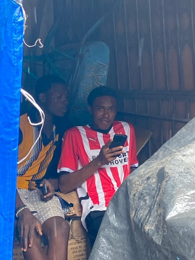 The image shows two young men in what appears to be a crowded, makeshift shelter. The man in the foreground, wearing a red and white striped shirt with "SPORT HOVE" visible on it, is holding a smartphone and looking at it. He has a relaxed posture. Partially visible behind him, another young man wears a yellow and black striped shirt. The shelter is covered with what looks like a large tarp or plastic sheeting, creating a dimly lit environment. Boxes are stacked in the background, some with visible text labels in English, suggesting a possible storage or temporary living situation. A blue tarp is visible in the foreground, suggesting an outdoor or partially exposed setting. The overall impression is one of everyday life in a potentially challenging environment, with the subjects finding moments of leisure or connection through their phones. The text "Ogbunike, Nigeria" in the prompt indicates the geographical context of this scene.