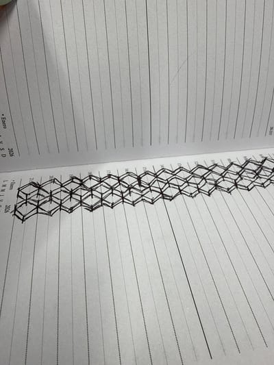 This image captures an open physical planner or diary, displaying two facing pages with a slight tilt. The pages are off-white and feature horizontal lines. On the right page, a vertical column of hourly markings (e.g., 9:00, 10:00, 11:00, 13:00, 14:00, 15:00, 16:00, 17:00, 18:00, 19:00, 20:00, 21:00) is visible along the left margin, indicating a daily or weekly schedule layout.
A prominent, intricate pattern, hand-drawn in black ink, runs vertically down the center of the open book, primarily on the right page. This elaborate design consists of interconnected diamond and zigzag shapes, giving it a woven or braided appearance. The drawing spans most of the visible hourly entries, suggesting a creative or decorative element added to the planner.
The left page is less structured, mainly showing horizontal dotted lines. The right page also features dotted lines for the schedule, with some thicker, solid lines cutting across them. Visible text includes "28-337" at the top left corner. At the bottom of both pages, "Enero 2026" is clearly legible, with "L M M J V S D" (likely representing days of the week: Lunes, Martes, Miércoles, Jueves, Viernes, Sábado, Domingo) also present on the right page, further confirming a calendar or planner context. The image illustrates the act of personal planning, journaling, or creative doodling within a physical medium, highlighted by soft, even lighting that shows the paper's texture.