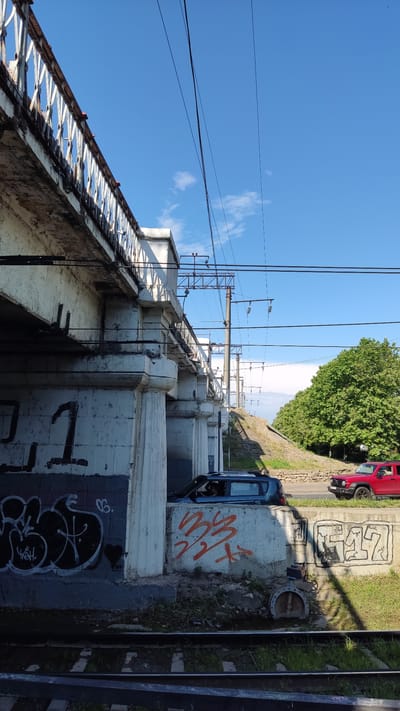 The image shows a bright, sunny day in Krasnodar, Russia. The scene is an elevated railway or overpass, characterized by its white concrete structure with decorative railings and a textured, peeling facade. A series of electrical wires and support structures for the railway are visible overhead, extending into the distance. Beneath the overpass, there are patches of grass and some debris.

On the street adjacent to the overpass, a red car is parked, and further down, a dark blue car is partially visible. Both vehicles appear to be stationary. The concrete supports of the overpass are covered in graffiti. One section of the concrete wall features bold orange lettering, while another section displays intricate black graffiti with white highlights.

The sky is a clear, vibrant blue with a few wispy white clouds. The sunlight casts distinct shadows, suggesting it is likely daytime, possibly late morning or early afternoon. The overall impression is an urban landscape with elements of infrastructure, transportation, and street art.