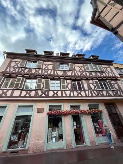 The image shows the facade of a building in Colmar, France, with typical Alsatian half-timbered architecture. The building has a pink stucco exterior with dark brown timber framing. There are several rows of windows with wooden shutters on the upper floors. The ground floor features two storefronts with large display windows. The left storefront has a sign that reads "Inspiration Design" and displays various items within. The right storefront has a sign that reads "Jade & Alize" and also displays clothing and accessories. A woman in blue jeans and a brown jacket is walking away from the right storefront. A flower arrangement adorns the top of the storefront windows. The sky above is blue with white, fluffy clouds.
