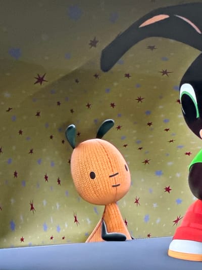 A partial view of a cartoon scene shows an orange, oval-shaped character with knit-like texture, small black dot eyes, and a short, wide neck. It has teal-colored limbs. Above it, a black, curved figure with tan markings and a bright green, crescent-shaped highlight is visible, as well as a small portion of another figure wearing red and white. The backdrop is a yellow-green color with scattered red star-shaped designs and small blue dots. A solid gray edge is on the right side.