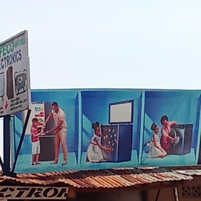 The image depicts an advertisement for FECO Electronics in Lafia, Nigeria. The advertisement features painted scenes of families interacting with appliances: a man and child at a refrigerator, two girls at a television, and two girls at a washing machine. The advertisement is painted on a large sign above a corrugated metal roof. Another sign for FECO Electronics stands to the left of the painted advertisement.
