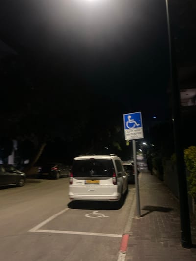 A white van parks in a designated handicap parking space at night. The handicap symbol is painted on the ground and a sign with the wheelchair symbol is posted. Other cars are parked nearby, and a street light illuminates the scene. The road and curb are clearly visible. The setting appears to be urban, with buildings and trees visible in the background.