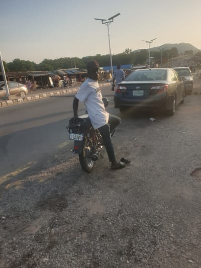 A man on a motorcycle with the license plate FST-520-EA is on a paved street in Zango-ndaji, Nigeria. A dark blue Toyota Camry is in the background. Other vehicles, buildings, and roadside stalls are visible on the side of the street. Streetlights are present. The terrain in the background includes a hill. The shoulder of the road consists of asphalt gravel.