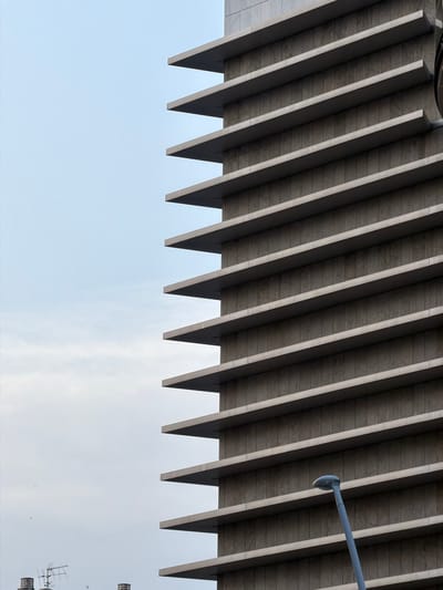 The image captures a close-up, low-angle shot of a modern building facade with horizontal protrusions, possibly balconies or architectural elements, creating a stepped effect. The building, made of light-colored stone or concrete panels, dominates the right side of the frame. The sky, a pale blue with subtle cloud textures, fills the left two-thirds of the image. A grey streetlamp with a rounded head extends upwards from the bottom right corner, angled towards the building. In the far distance, on the left, faint outlines of other buildings and what appear to be rooftop antennas are visible. The overall scene suggests an urban environment, likely in the city of Castelló de la Plana, Spain, given the architectural style and common urban elements. There are no people or overt actions depicted; the focus is on the geometric forms of the architecture and the sky.