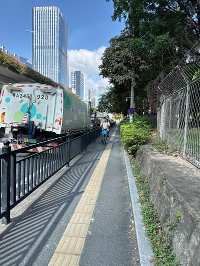 A cyclist rides a blue bicycle on a paved pathway in the Huangpu District, China. The pathway is bordered by a black metal fence on the left and a concrete embankment with vegetation on the right. In the background, a white garbage truck with colorful circular designs is parked on the adjacent road. Beyond the truck, modern high-rise buildings with reflective glass facades dominate the cityscape. The sky is clear with scattered white clouds. A street light and a traffic sign are visible along the pathway. The scene is illuminated by sunlight, casting shadows from the fence onto the pathway.
