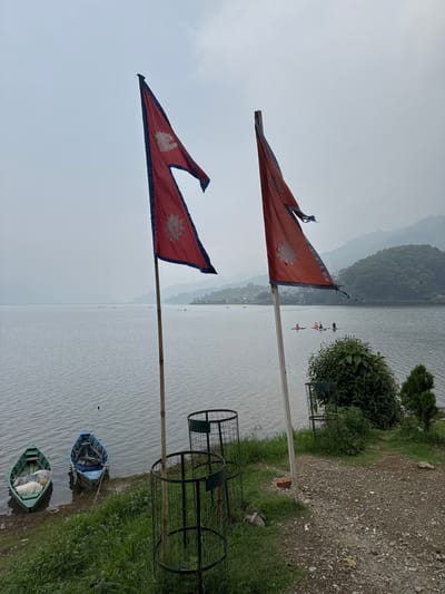 This photograph shows two flags of Nepal fluttering from poles on a grassy bank next to Fewa Lake in Pokhara, Nepal. Two metal cage-like trash cans are visible near the edge of the bank and two small boats, one blue and one green, sit in the water near the grass. In the distance, there are people in kayaks and mountains. The sky is overcast.