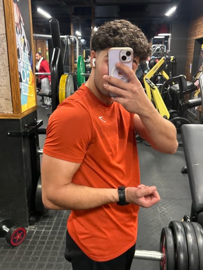 A man in a gym setting is taking a selfie. He is wearing an orange shirt and black shorts. He holds a phone in his left hand. His right arm is flexed to show off his bicep. There is gym equipment in the background, including weight machines and dumbbells. The floor has a gray, textured surface. A poster is visible along the bottom edge of the frame. The light in the gym is bright.