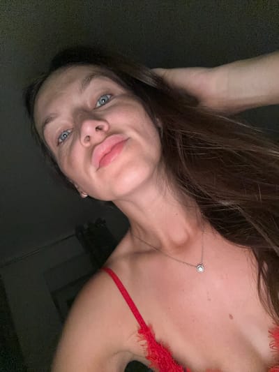 In an indoor setting, possibly a bedroom in Donji Pobori, Montenegro, a young woman with long, dark brown hair is captured in a selfie. She is looking directly at the camera, her head tilted slightly. Her piercing blue eyes are noticeable, and her lips are pursed in a playful expression. A delicate necklace with a small, circular pendant rests on her collarbone, adding a subtle touch of elegance. She is wearing a vibrant red top with lace detailing, hinting at intimate apparel. The lighting is dim, with shadows obscuring much of the background, creating an intimate and personal atmosphere. Her left hand is raised, fingers brushing through her hair, suggesting a relaxed and casual pose. The image is framed from the chest up, focusing on her facial features and upper body.