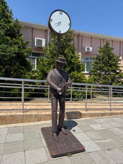 In Slavyansk-na-Kubani, Russia, a bronze statue of a man in a suit and hat stands on a dark rectangular base. The statue is positioned outdoors on a paved walkway, with a building in the background. The building has a light pink facade, white trim, and several windows. Green trees and bushes are visible around the statue and in front of the building. Above the statue, a large clock with Roman numerals and thin black hands is mounted on a pole. The clock face is white with a dark circular rim. The sky is clear and blue. A metal railing runs horizontally behind the statue, separating it from the building. The ground is paved with light-colored square tiles with some dark grout lines. The statue appears to be looking down at its hands, as if checking a watch.
