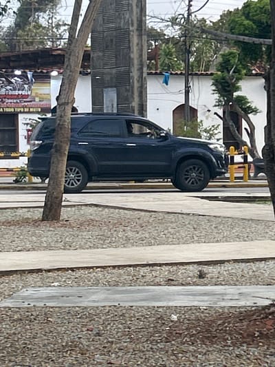 The image shows a dark blue SUV parked on a street in Santa Ana, Venezuela. The vehicle is positioned in the foreground, with its side facing the viewer. The surrounding environment includes trees, buildings, and a rocky, gravel-covered area. A wooden pole with a blue cloth draped over it is visible in the background. The sky appears overcast.