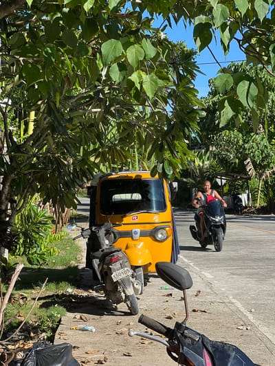 This is a ground-level, eye-level, full shot taken in General Luna, Philippines, on a bright, sunny day. The scene is dominated by lush green foliage from trees overhead, casting dappled shadows on the ground. In the foreground, a black motorcycle is parked, with its handlebars and side mirror visible. To the left, a bright yellow auto-rickshaw, or "tricycle" as it's known locally, is parked, with its front wheel and lights visible. Behind the auto-rickshaw, a grey scooter is parked, displaying the license plate "150105". Further down the paved road, a man is riding a black scooter. He is bald, wearing a dark sleeveless shirt and red shorts, and is looking towards the camera. The road is lined with vegetation and a few small structures, including a covered stall on the right side of the road. The environment appears tropical, with clear blue skies visible through the leaves.
