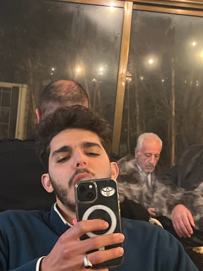 A selfie is taken in a mirror reflection. The person in the foreground holds a cell phone with a ring accessory on its back. They wear a silver ring and a navy sweater. Reflections in the background show two other people, including one with white hair holding something that appears to be smoking. The surroundings look like a restaurant or bar, with a window and lights visible in the reflection.
