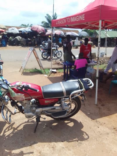 This image depicts a bustling outdoor scene in Ukwu-Abwa, Nigeria, under bright daylight conditions. In the immediate foreground, a red motorcycle with a black seat, chrome accents, and a rear rack holding a dark item is parked on a dirt surface, supported by its kickstand. To the right of the motorcycle, a red pop-up canopy tent, bearing the text "A REASON TO IMAGINE," is set up. Under this tent, a man in a red "Airtel" branded t-shirt stands behind a table, facing left. He appears to be interacting with a woman wearing a bright pink top, seated at the table with her back to the camera. Another man in a dark sleeveless top stands to the left of the table. A fourth individual, wearing striped trousers, is partially visible seated further right.
A dirt path and an A-frame signboard separate this activity from a paved road in the midground. On the road, two motorcycles with riders are visible, indicating traffic. Beyond the road, the background features a dense collection of market stalls, covered by various tarpaulins and umbrellas in muted colors, suggesting a vibrant market environment. Numerous indistinct figures move among these stalls. The sky above is partly cloudy, and sparse vegetation is visible in the distance.