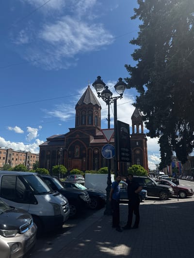 The image depicts a view of the Saint Mary of God Cathedral in Gyumri, Armenia, on a bright, partly cloudy day. The cathedral, constructed from dark red and black stone with decorative orange trim, is the central architectural feature, characterized by its prominent conical roof and bell towers. In the foreground, a row of parked cars lines a street, including a white transit van, a dark SUV, and a dark sedan. Two men are standing on the sidewalk near a lamppost, with one leaning against it. A street sign with directional arrows is also visible on the lamppost, alongside traffic signs. Residential buildings with a reddish-orange facade are seen in the background to the left of the cathedral, and a large conifer tree with dense foliage partially obscures the right side of the scene. The sky is a vivid blue with scattered white clouds, and power lines are strung across the upper portion of the frame. The overall setting appears to be a public area with a mix of historical and modern elements.