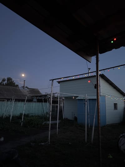 The image depicts an outdoor scene at dusk or twilight, likely in Rodino, Russia. The sky is a deep blue, suggesting the evening or early night. A string of colorful lights is visible, draped across a metal pole that runs horizontally across the upper portion of the frame. These lights are a mix of red, yellow, green, and blue, adding a festive or decorative element to the scene.
In the background, a light blue house with white trim is partially visible. It has a small window near the peak of its roof and what appears to be blue-tinted windows or doors on a lower level. Adjacent to this house is another building with a corrugated metal roof. A chimney rises from this structure.
A notable detail in the sky is the moon, a bright orb visible in the upper left quadrant of the image, partially obscured by clouds. Trees can be seen on the left side, silhouetted against the darkening sky.
The foreground is dominated by the dark, angled roof of another structure, casting a significant shadow and obscuring details beneath it. The ground appears to be covered in grass and dirt, with some areas darker and more shadowed than others. There are some white poles or supports visible, suggesting some kind of outdoor structure or perhaps clotheslines. The overall atmosphere is quiet and serene, with the glow of the moon and the string lights suggesting a peaceful evening. There is no visible text in the image.