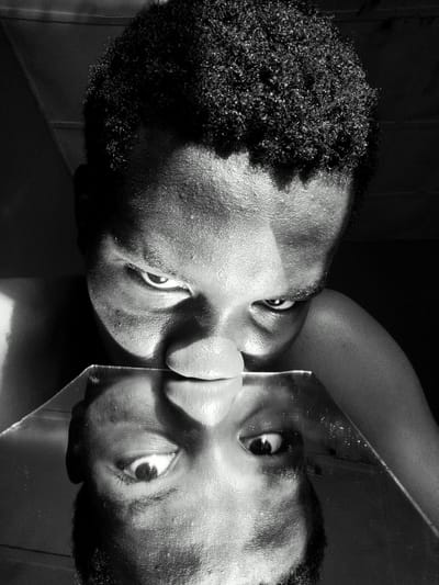 A black and white close-up photograph features a dark-skinned person with curly hair looking downward into a small mirror held at nose level. The face reflected in the mirror is an upside-down version of the same person. The scene gives the impression of being indoors, with shadows in the background. There is no clear indication that it was taken in the city of Keffi, Nigeria.