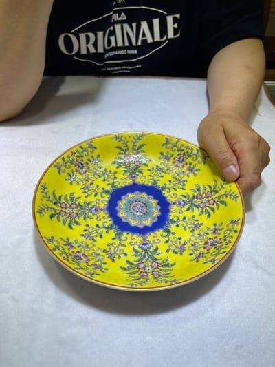 A hand holds a yellow ceramic plate with a floral pattern. The plate is decorated with intricate designs of flowers, leaves, and swirling vines in shades of pink, purple, green, and blue. A central medallion features a complex geometric and floral motif in blue and green. The plate has a thin red and gold rim. The background is a white, textured surface, possibly a tablecloth or fabric. To the left of the plate, a person's arm and shoulder are visible, wearing a black t-shirt with white text that reads "FILA ORIGINAL". The text is in a large, bold font. The scene appears to be indoors, likely a studio or home setting, given the controlled lighting and plain background. The lighting suggests it is daytime. The overall impression is of a close-up view of an ornate decorative object.