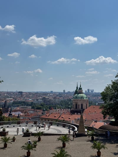 This is an elevated, daytime, full shot of a city skyline in Prague, Czechia, viewed from a terrace. The sky is a bright blue with scattered white cumulus clouds. In the foreground, a paved terrace area is furnished with potted palm trees and a large, pyramid-shaped stone monument. Several people are walking on the terrace, some holding umbrellas for shade. A curved walkway with a railing separates the foreground terrace from a lower level where more people are walking.
Beyond the terrace, the city unfolds with numerous red-tiled roofs and buildings. A prominent green-domed church with two spires stands out in the middle ground, identified as St. Nicholas Church in Malá Strana. In the far distance, modern high-rise buildings punctuate the historical cityscape. Trees are interspersed throughout the urban landscape, and the Vltava River is visible as a sliver in the distance. The overall scene depicts a sunny day in a historic European city.