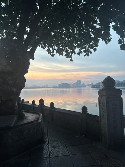 The image captures a serene waterscape at dawn or dusk, with a city skyline faintly visible in the distance across the water. The sky displays a gradient of soft blues and warm yellows, indicative of sunrise or sunset. In the foreground, a large tree silhouette dominates the left side, its dark leaves contrasting with the lighter sky. On the right, a stone structure, likely part of a temple or monument, features intricate carvings and decorative finials. The water is calm, reflecting the colors of the sky and the distant city lights. The overall mood is peaceful and contemplative, evoking a sense of tranquility. The scene is set in Hà Nội, Vietnam, suggesting a location near a lake or river within the city. No people are visible, and no text is discernible in the image.