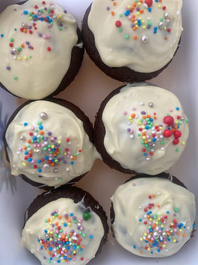 The image displays a top-down view of six chocolate cupcakes arranged in a white rectangular container. Each cupcake is topped with white frosting and adorned with a scattering of colorful sprinkles and silver edible pearls. The cupcakes appear to be homemade, with the frosting applied unevenly, creating a rustic aesthetic. There are no people or actions visible in the image. The environment is a close-up shot of the desserts, with no discernible background elements to indicate a specific location within Podgorica, Montenegro, or any other city.