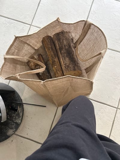 The image, captured in Veauchette, France, presents a high-angle view of an indoor scene. The lower portion of a person, identifiable by legs clad in black sweatpants, occupies the bottom right quadrant. Adjacent to the person, a beige burlap bag stands open, containing multiple cut firewood logs of varying sizes and bark textures. The bag and person are positioned on a light-colored tiled floor with a visible grid pattern. To the left, a dark, circular metallic base of an object, possibly a stove or fireplace component, is partially visible, surrounded by a light dusting of ash and debris. A thin, black, rod-like object rests horizontally on the tiled floor near the metallic base.