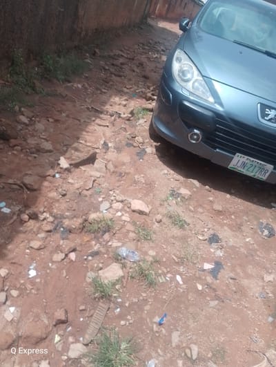 The image captures a close-up view of a grey Peugeot 307 parked on a dirt road in Gura Top, Nigeria. The car's front is angled towards the right side of the frame, revealing its headlights, grille, and license plate, which reads "JJN-217RR". The road surface is uneven, composed of dirt, small rocks, and scattered debris, including plastic wrappers and what appears to be a broken plastic bottle. Patches of dry grass grow sporadically amidst the dirt. A textured, reddish-brown wall runs along the left edge of the image, suggesting the car is parked near a building. The overall scene is unkempt, indicating a neglected or rural environment. No people are visible in the image.