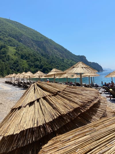 A sunny day at a beach in Prijevor, Montenegro. In the foreground, several straw umbrellas are stacked, with their bamboo poles visible. Beyond them, a row of open straw umbrellas stands over empty lounge chairs. The beach is covered in pebbles and sand. In the background, the turquoise sea is visible, with a few people sunbathing on loungers and swimming. A green, forested mountain slopes down towards the sea. The sky is clear and blue.