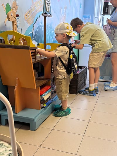 Two boys and an adult are in a room with a mural on the wall depicting a ship and a fairy. A younger boy, wearing a Pikachu-themed hat, shorts, and a backpack, is playing with a toy cash register. He is wearing green shoe covers. An older boy, wearing a light green t-shirt and shorts, is standing next to him, looking at something on the cash register. He is wearing blue and yellow shoes. The adult, wearing a blue top and a denim skirt, is standing to the right, holding a lavender bag. The room has tiled floors and a wooden chest is visible behind the younger boy. This scene is located in Podolsk, Russia.