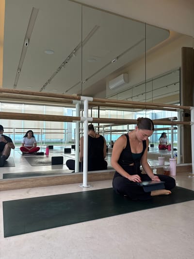 In San Bernardino Tlaxcalancingo, Mexico, a yoga class is taking place in a studio with a large mirrored wall. In the foreground, a woman in black athletic wear sits cross-legged on a dark yoga mat, focused on a tablet. Behind her, reflected in the mirror, are other individuals participating in the class, including a woman in a white top and red leggings and another in a black tank top. The studio features wooden barre equipment along the mirrored wall, and large windows in the background offer a view of the outdoors. The floor is a light, polished concrete.