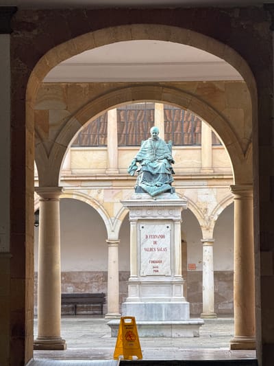 The image depicts a statue of D. Fernando de Valdés Salas, the founder of the University of Oviedo in Spain, as seen through an archway. The statue stands on a marble plinth, with an inscription indicating his name and the founding dates of 1608 and 1908. The plinth is located in a courtyard or interior space with classical architectural elements, including columns and additional arches. A yellow "Caution Wet Floor" sign is visible to the right of the statue. The architectural style suggests a historic building, consistent with the university's age. The image appears to be taken from within the building, framing the statue through an opening.