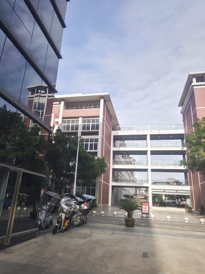 The image depicts an outdoor scene in the city of Chongqing High-tech Zone, China, featuring modern architecture and some parked vehicles. In the foreground, to the left, is a portion of a large, contemporary building with dark, reflective glass windows. Adjacent to this building, on the pavement, are three electric scooters, each covered with a clear plastic canopy. Two of the scooters are parked closer together, while the third is slightly behind and to the right.

The background reveals another substantial building complex constructed with red brick and white accents. This building has multiple levels, including elevated walkways with glass railings connecting different sections. Trees with green foliage are visible around the buildings, suggesting a well-maintained urban environment. The sky above is partly cloudy, indicating a daytime setting. A sign with Chinese characters and "No parking FIRE LANE" in English is positioned near a potted plant in the middle ground. Several white, spherical bollards line the paved area in front of the buildings. The overall impression is one of a modern educational or administrative campus.