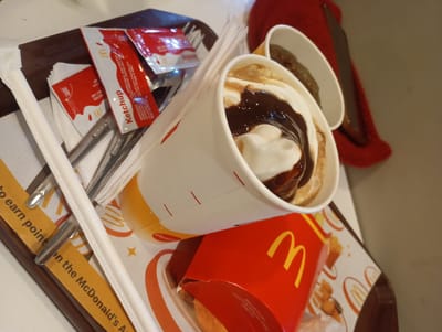 The image shows a close-up of a McDonald's meal served on a tray. The primary subjects are food items and packaging from McDonald's. These include a white cup with a dessert, likely an ice cream sundae with chocolate sauce and whipped cream, a red McDonald's french fry box, and several packets of ketchup and what appears to be salt. A white paper straw and some cutlery are also visible. The setting is a McDonald's restaurant, suggested by the branded packaging and the design of the tray liner which features the McDonald's logo and images of french fries and chicken nuggets. The lighting suggests it is daytime indoors. There is text visible on the ketchup packets, including "Ketchup" and "McDonald's," and on a yellow paper strip on the tray that says "to earn point on the McDonald's A". No people are visible in the frame. The overall impression is of a casual meal being enjoyed.