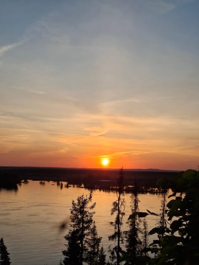 The image is a landscape-oriented photograph of a sunset over a body of water, possibly taken near Ольховка, Russia. The sun is setting on the horizon, casting an orange and yellow glow across the sky and reflecting in the water. The sky has hues of blue on the left side. Silhouetted trees and foliage are visible on the right side of the frame. Some trees are mirrored in the water. The water appears still.
