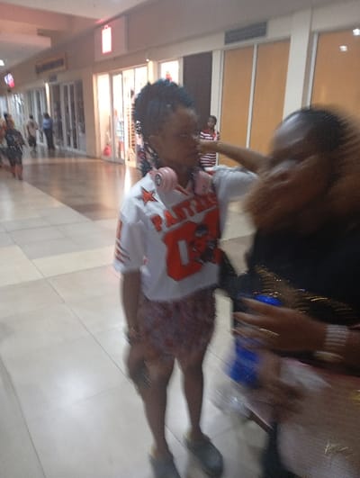 A young woman in a Panthers jersey, patterned shorts, and slippers is standing in a mall. She has pink headphones around her neck. A person with curly hair is partially in the frame to her right. Several shops line the mall's sides, and people walk in the distance. The floor is tiled.
