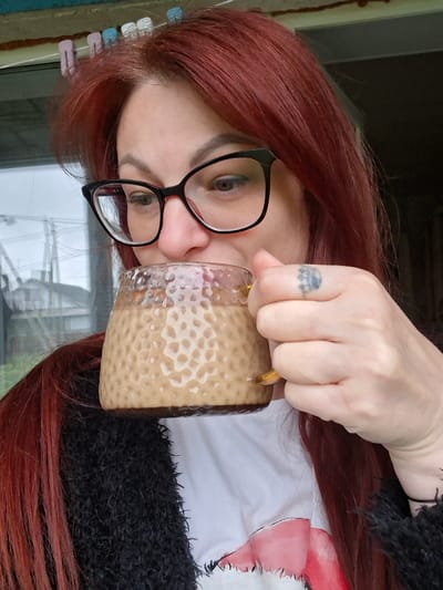 In a close-up, upper body shot, a woman with long, dark red hair and black-rimmed glasses is holding a textured glass mug with a light brown beverage up to her mouth, as if drinking. She is wearing a black, fuzzy jacket over a white t-shirt with a red graphic design. A small, crown-shaped tattoo is visible on the back of her right index finger. The background shows a blurry outdoor scene with what appears to be houses and a cloudy sky, suggesting a residential area. This scene is likely located in or around Забалоцкі сельскі Савет, Belarus.